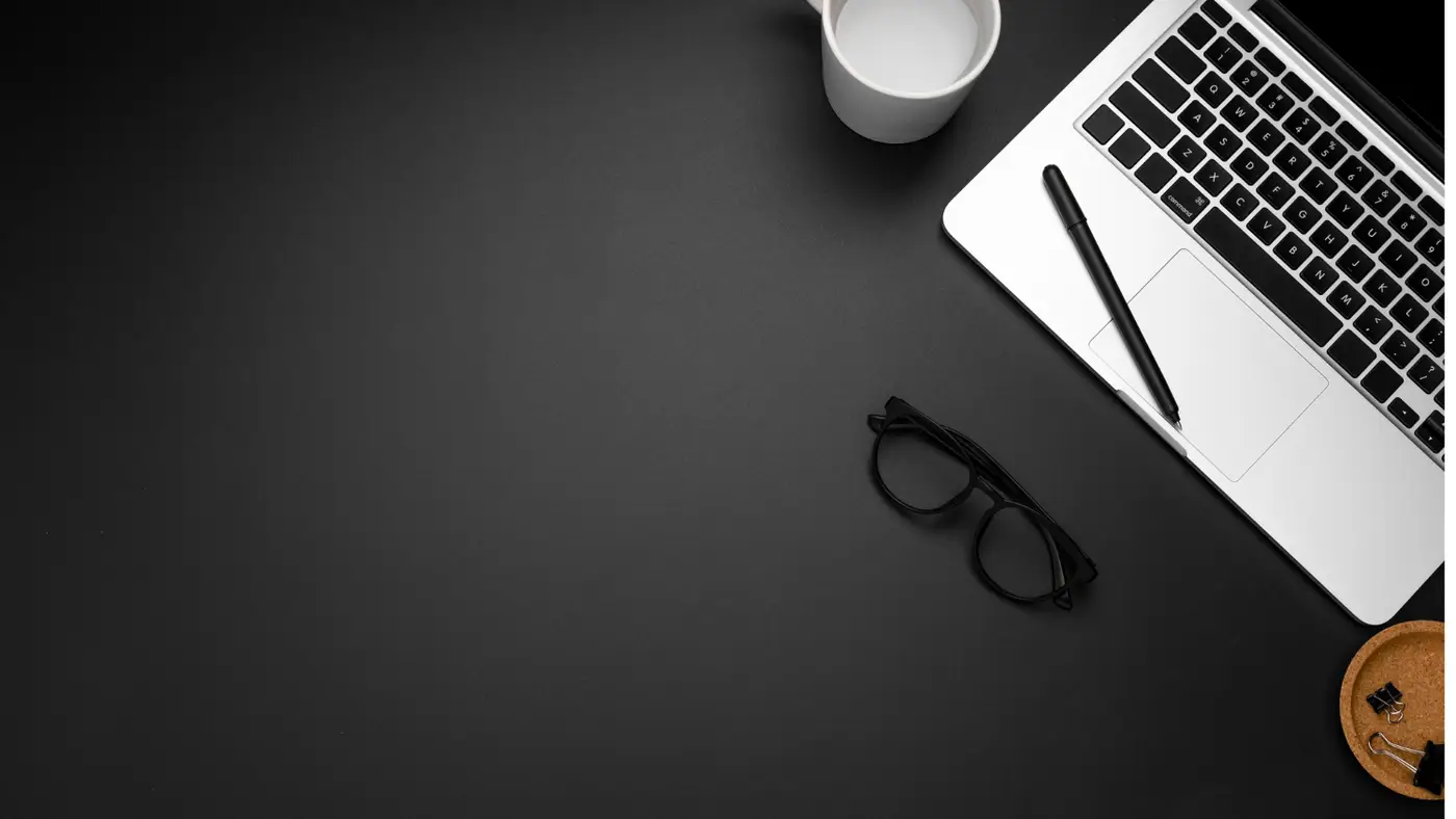A laptop, a pen and reading glasses on a dark desk.